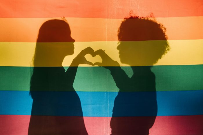 Pareja de mujeres formando un corazón con las manos frente a una bandera LGBTQIA+